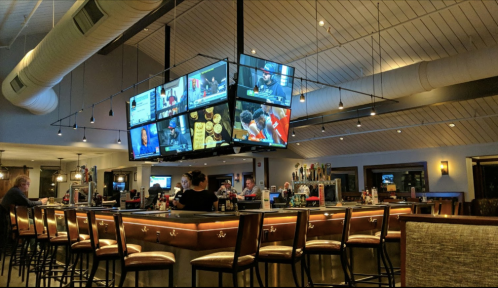 A modern bar with multiple TVs displaying sports, patrons seated at the counter, and a warm, inviting atmosphere.