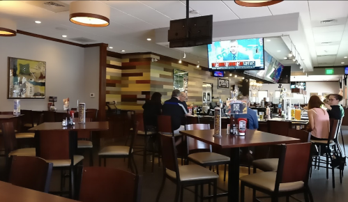 A casual restaurant interior with wooden tables, a bar area, and a TV showing a sports game. Patrons are seated at tables.