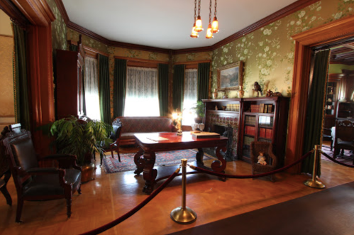 A vintage room with floral wallpaper, a wooden table, chairs, and a cozy seating area, illuminated by warm light.