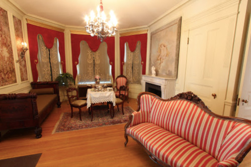 A vintage parlor with a striped sofa, elegant chandelier, and ornate decor, featuring a small table and decorative curtains.