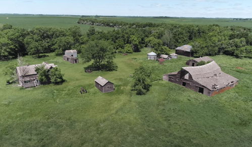 Aerial view of an abandoned farmstead with several old buildings surrounded by lush green fields and trees.