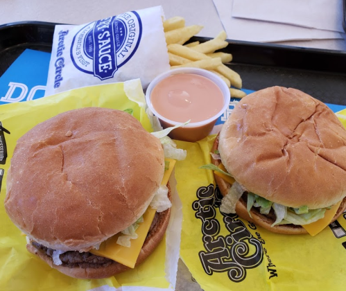 Two burgers with cheese and lettuce on yellow wrappers, accompanied by a side of fries and a dipping sauce.