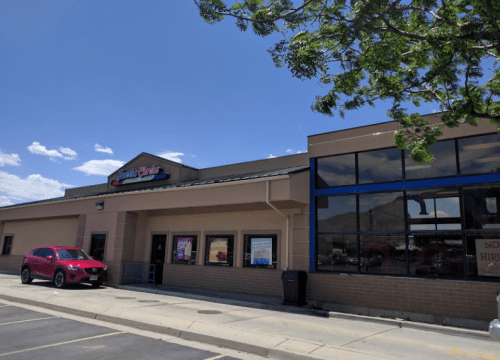 Exterior of a restaurant with a blue and beige facade, large windows, and a pink car parked outside. Clear blue sky above.