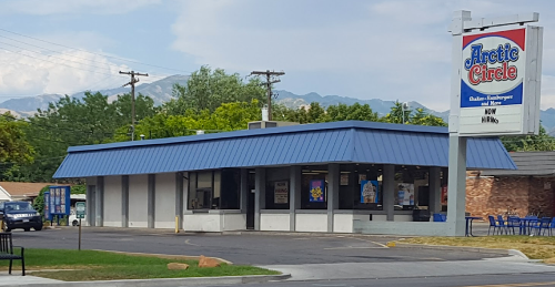 A fast-food restaurant with a blue roof and sign, surrounded by trees and mountains in the background.