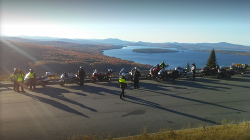 A group of motorcyclists stops to admire a scenic view of a lake surrounded by autumn foliage.