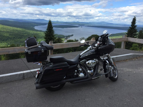 A black motorcycle parked on a scenic overlook with a lake and mountains in the background under a cloudy sky.