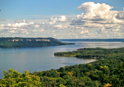 A scenic view of a river surrounded by lush greenery and rolling hills under a partly cloudy sky.