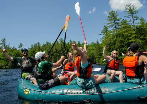 A group of six people in life jackets celebrate on a raft, paddles raised, surrounded by trees and a clear blue sky.