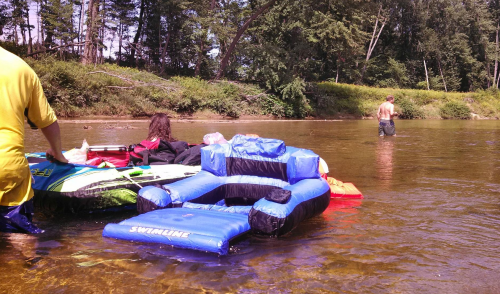 Two people are near inflatable rafts in a shallow river, with one person wading in the water and trees in the background.