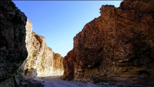 A narrow canyon with towering rocky walls under a clear blue sky. Sunlight illuminates the rugged terrain.