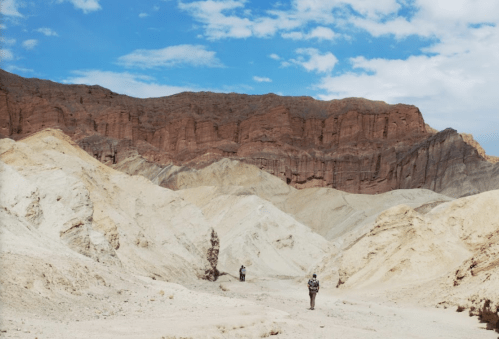 Two hikers explore a colorful, rugged landscape of layered rock formations under a blue sky with scattered clouds.