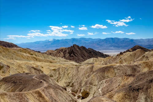 A vast desert landscape with rugged, multicolored mountains under a bright blue sky with scattered clouds.