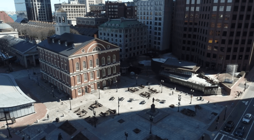 Aerial view of a historic building surrounded by modern architecture and a plaza with people and benches.