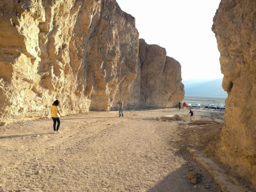 Two people walking in a rocky canyon with a distant view of parked cars and mountains under a clear sky.