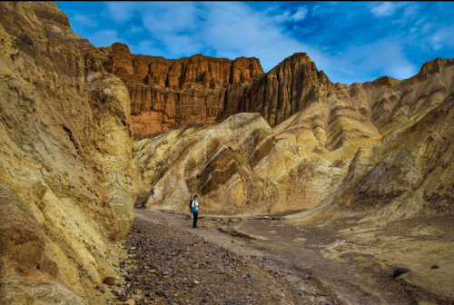 A person stands in a colorful, rugged canyon with steep, layered rock formations under a blue sky.