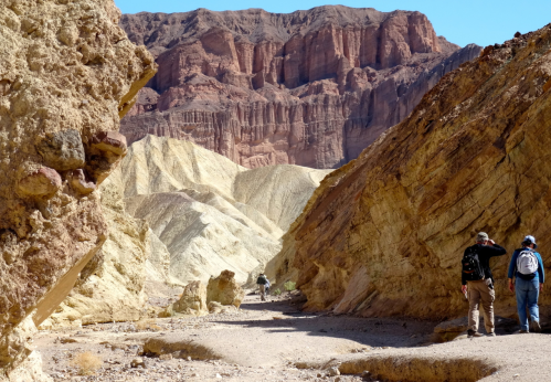Two hikers explore a narrow canyon surrounded by colorful rock formations and towering cliffs under a clear blue sky.