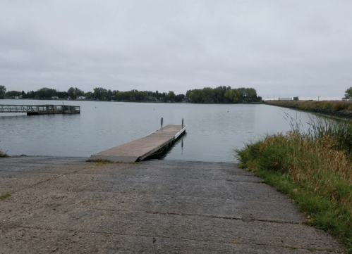 A wooden dock extends into a calm lake, surrounded by greenery under a cloudy sky.