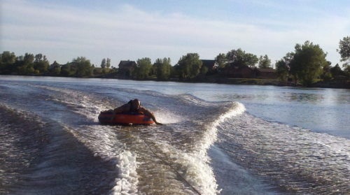 A person on an orange inflatable tube is being towed across a calm river, creating ripples in the water.