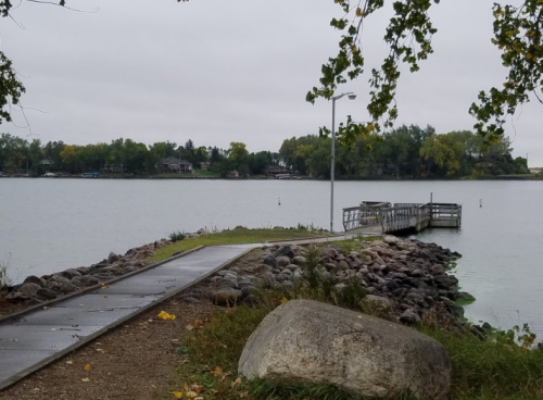 A calm lake scene with a wooden dock, surrounded by trees and rocky shoreline under a cloudy sky.