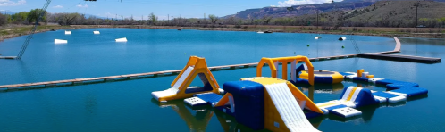 A colorful inflatable water park with slides and obstacles on a blue lake, surrounded by mountains and clear skies.