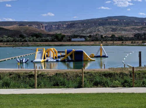 A colorful inflatable water park sits on a lake, surrounded by green grass and mountains under a blue sky.