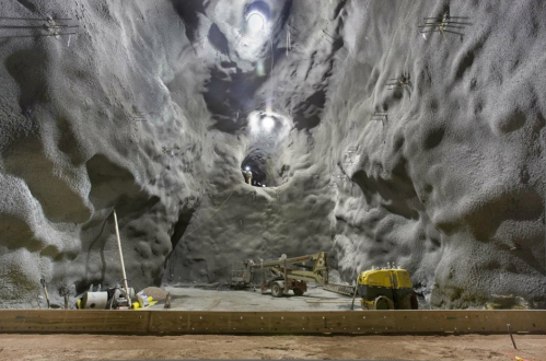 A large, cavernous underground space with textured walls, construction equipment, and overhead lights illuminating the area.