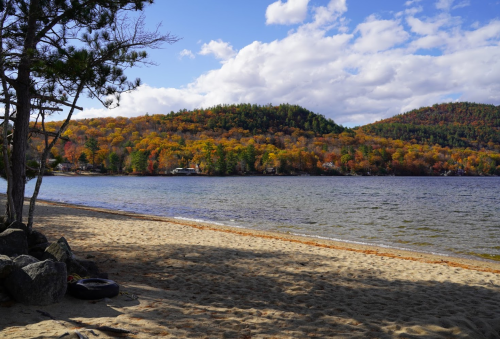 A serene lakeside view with sandy beach, colorful autumn trees, and rolling hills under a partly cloudy sky.
