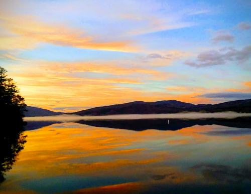 A serene lake at sunset, reflecting colorful skies and distant mountains, with a light mist hovering over the water.