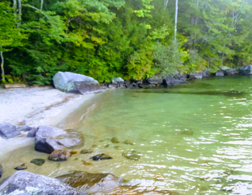 A serene lakeside scene with clear water, smooth rocks, and lush green trees lining the shore.