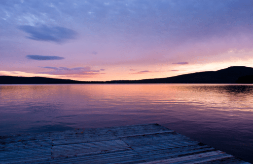 A serene lake at sunset, with vibrant pink and purple hues reflecting on the water and a silhouette of distant hills.