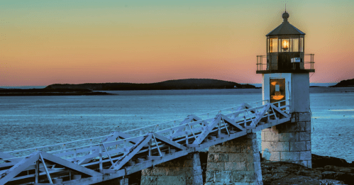 A lighthouse at sunset, with a wooden walkway leading to it over calm waters and distant hills.