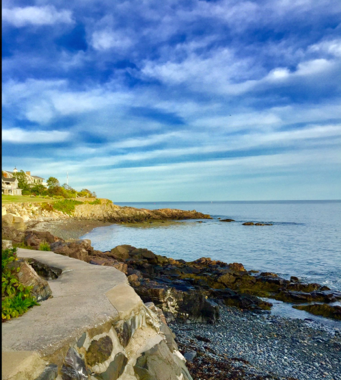 A scenic coastal view with rocky shores, calm water, and a blue sky dotted with clouds.