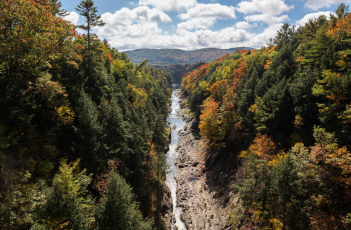 A scenic view of a river winding through a colorful forest in autumn, surrounded by mountains and blue skies.