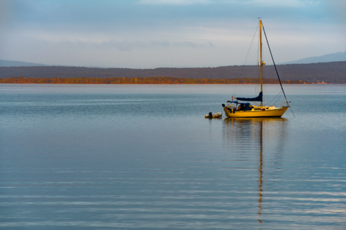 A calm lake at dawn with a yellow sailboat anchored, reflecting on the water's surface.