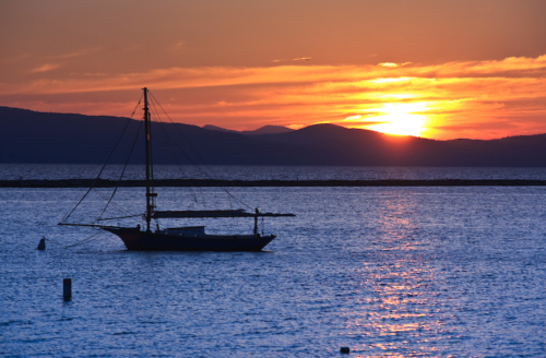 A sailboat anchored on calm waters at sunset, with mountains silhouetted against a colorful sky.