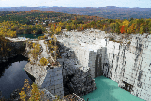 Aerial view of a quarry with turquoise water, surrounded by autumn foliage and distant mountains.