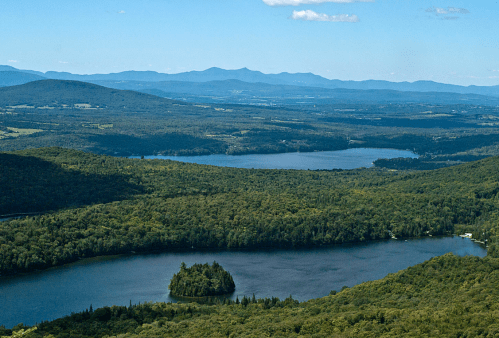 A panoramic view of lush green mountains and lakes under a clear blue sky, with an island in the foreground.