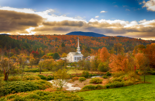 A scenic view of a white church surrounded by vibrant autumn foliage and mountains under a cloudy sky.