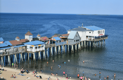 A wooden pier extends over calm blue waters, with beachgoers enjoying the sandy shore below.