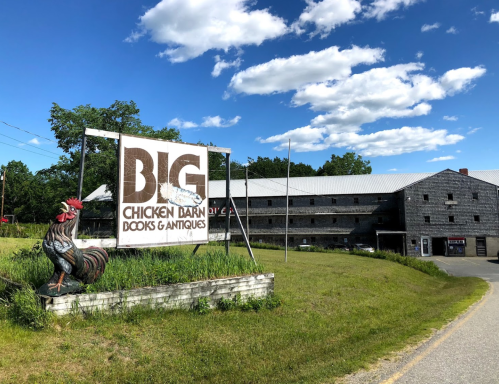 Sign for "Big Chicken Barn Books & Antiques" with a rooster statue in front of a large, rustic building under a blue sky.