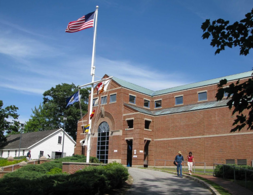A brick building with a flagpole displaying multiple flags, surrounded by greenery and two people walking nearby.
