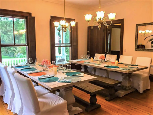 A bright dining room with a long table set for a meal, featuring white chairs, teal accents, and elegant lighting.