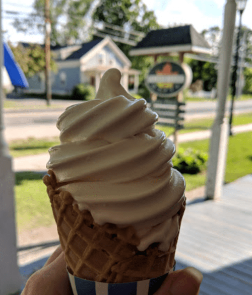 A hand holding a soft-serve ice cream cone with a swirl of creamy vanilla, set against a sunny outdoor background.