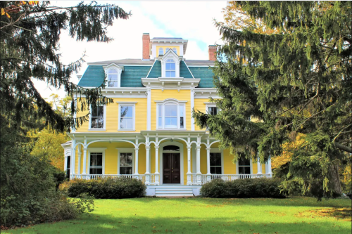 A large yellow Victorian house with green accents, surrounded by trees and a grassy lawn.