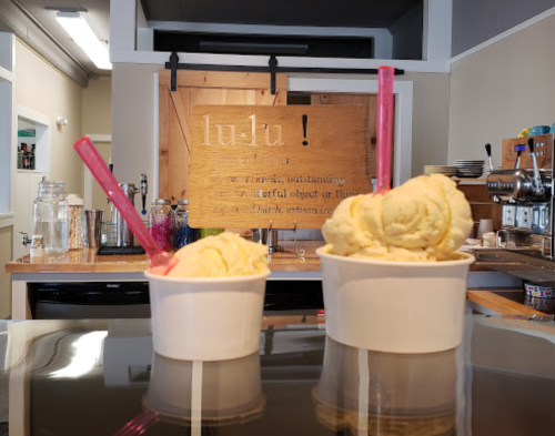 Two cups of ice cream with pink spoons sit on a counter, with a wooden sign reading "lu:lu!" in the background.