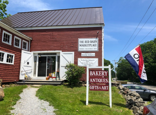 A red barn building with signs for a marketplace, bakery, antiques, gifts, and art, set in a rural area.