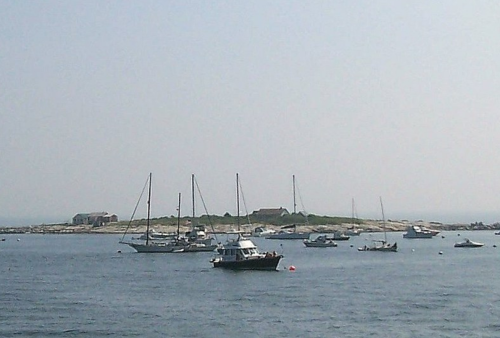 A serene harbor scene with several boats anchored near a small island and a distant shoreline under a hazy sky.