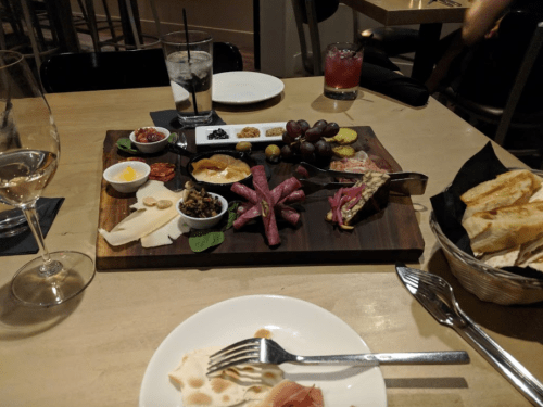 A charcuterie board with various meats, cheeses, olives, and accompaniments, alongside drinks and a basket of bread.