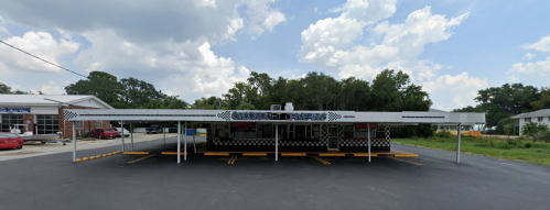 A retro-style diner with a checkered facade and outdoor seating under a canopy, surrounded by trees and a cloudy sky.