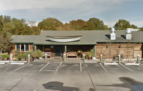A wooden building with a sloped roof, surrounded by trees, and a parking lot in front.